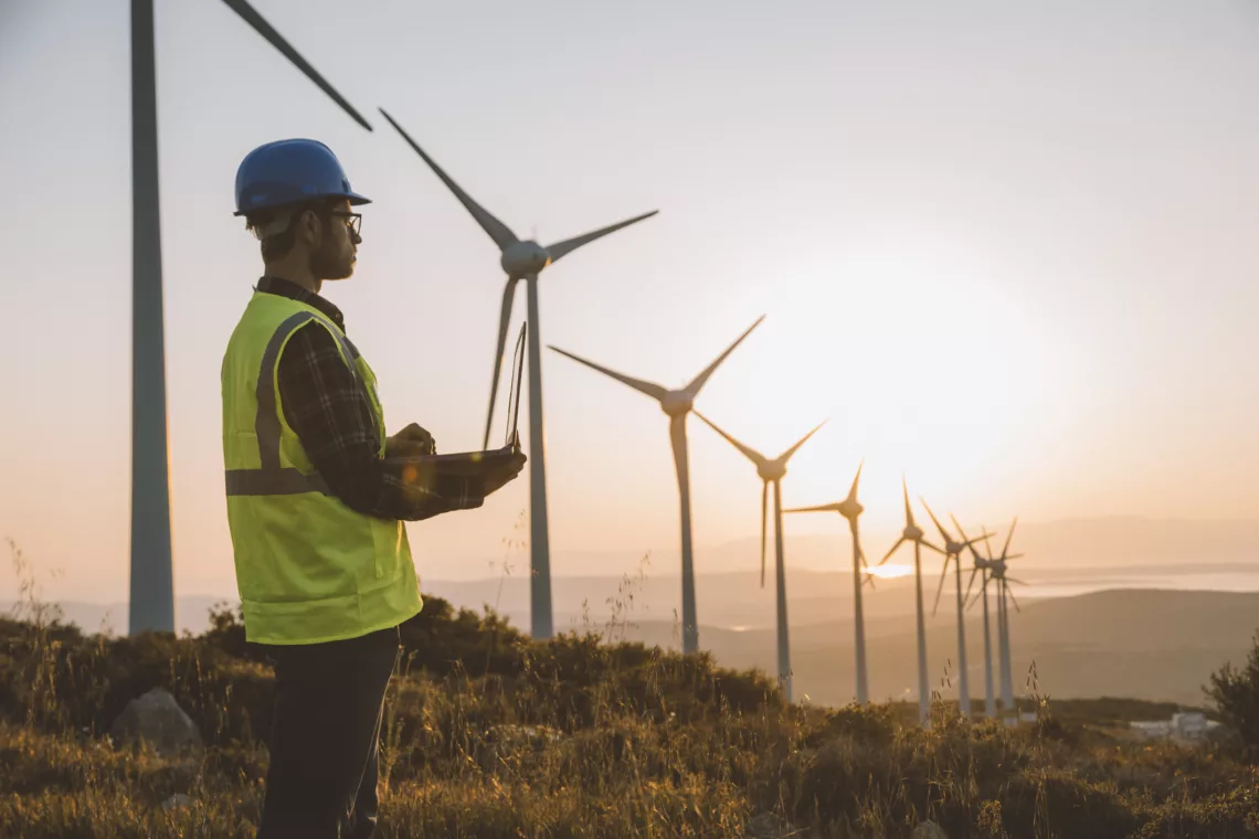 Silhouette of young male engineer holding laptop computer planning and working for the energy industry and standing beside a wind turbines farm power station at sunset time. XXXL size taken with Canon 5D MIV