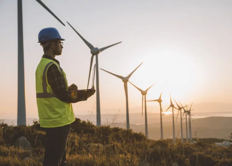 Silhouette of young male engineer holding laptop computer planning and working for the energy industry and standing beside a wind turbines farm power station at sunset time. XXXL size taken with Canon 5D MIV